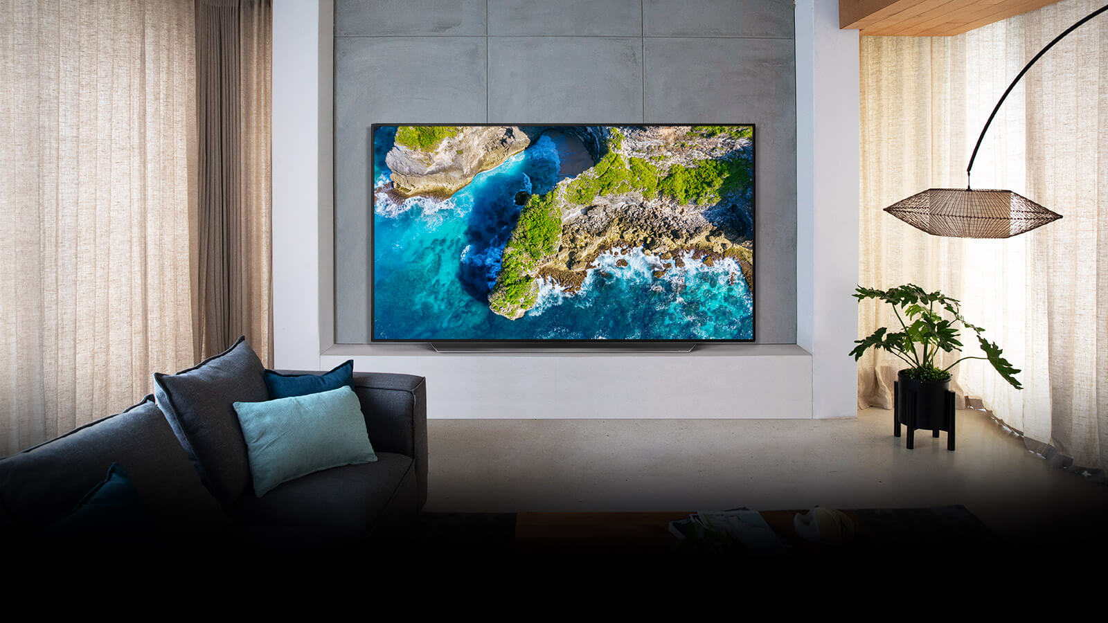 Ein OLED-TV hängt an einer grauen Wand in einem sehr modernen Wohnzimmer. Der Bildschirm zeigt eine Luftaufnahme von Meer und grün bewachsenen Klippen, um die Brillanz der Farben zu verdeutlichen.
