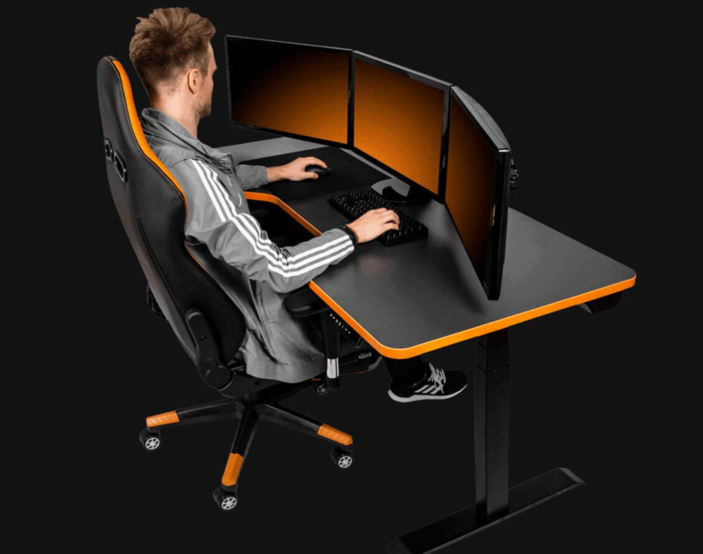 Ein Gamer sitzt an der schwarz-orangen Version eines Leetdesk Gaming-Schreibtischs und blickt auf drei Monitore. Der Hintergrund des Bildes ist komplett schwarz.