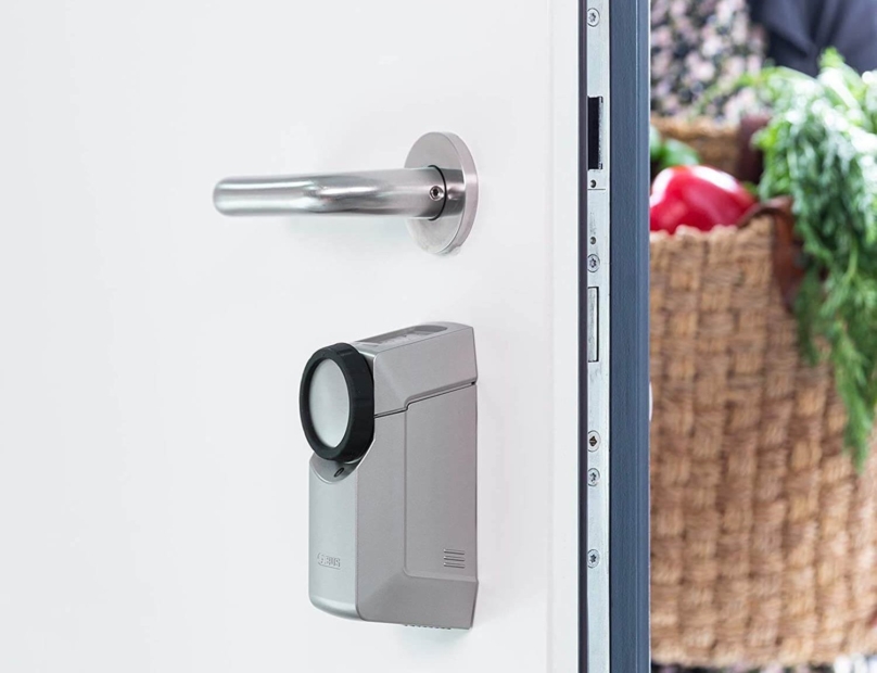Eine Tür mit Smart-Lock steht halb offen, während jemand mit einem Korb voller Einkäufen hereinkommt.