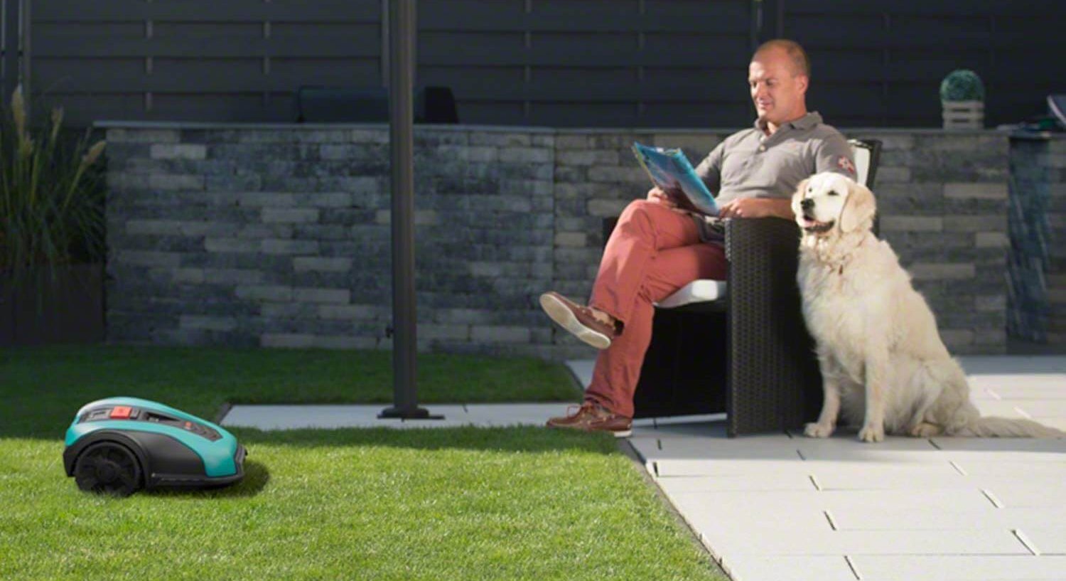 Ein Mann sitzt auf der Terrasse, neben ihm sein Golden Retriever. Auf der Wiese davor fährt ein Mähroboter von Gardena.