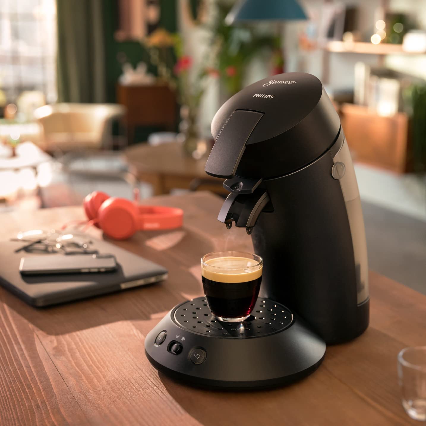 Eine Senseo Original Plus in der Farbe Schwarz steht auf einer hölzernen Küchentheke. Eine frische Tasse Kaffee dampft, im Hintergrund liegen ein Laptop, ein Handy und orange Kopfhörer.